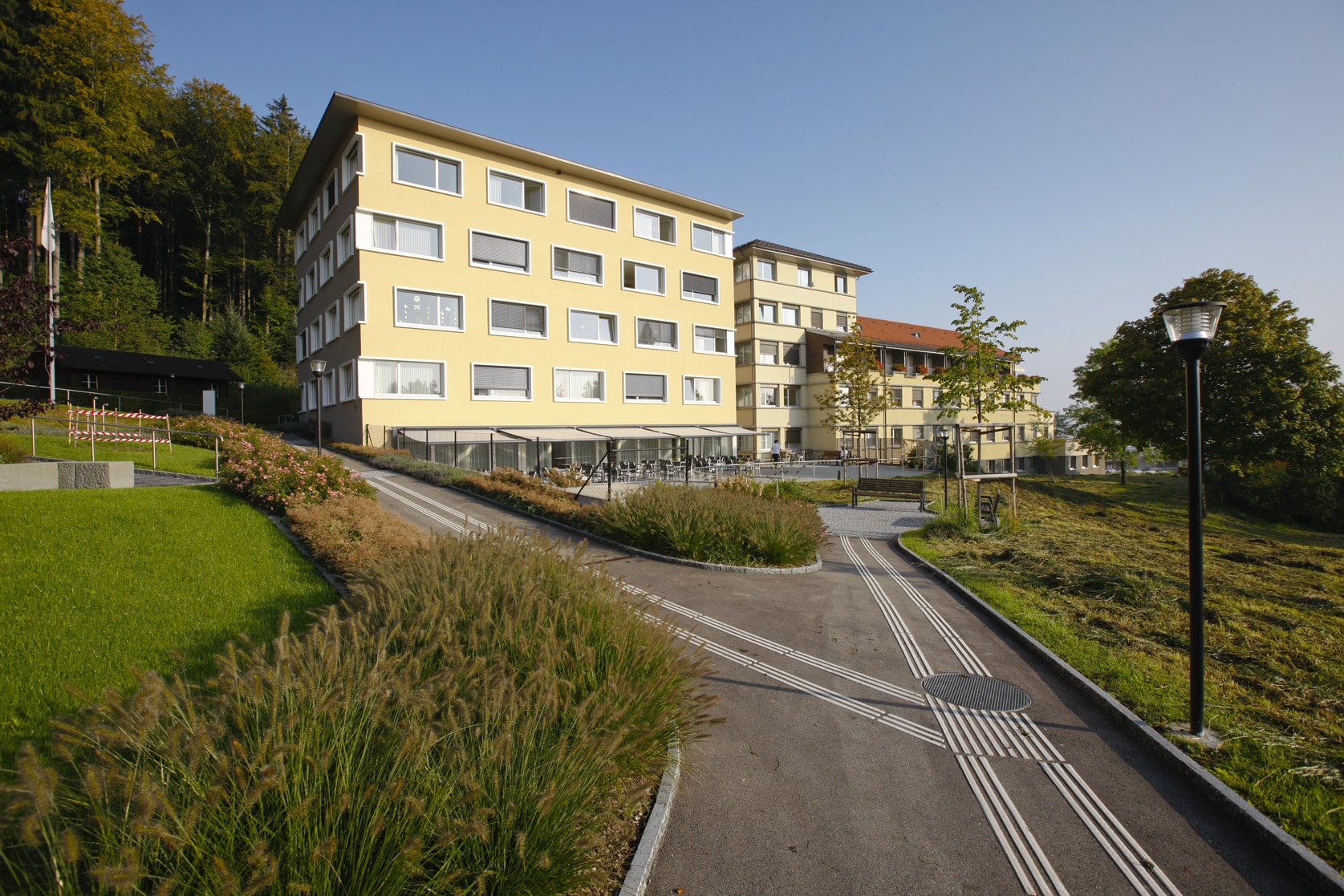 Ein modernes, hellgelbes mehrstöckiges Gebäude mit großen Fenstern, umgeben von Grün, Wegen und einem Laternenpfahl unter klarem Himmel.