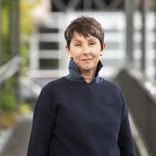 Eine Person mit kurzen braunen Haaren und einem marineblauen Pullover steht draußen auf einem Gehweg und blickt mit neutralem Gesichtsausdruck in die Kamera.