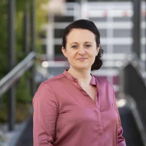 Eine Frau mit zurückgekämmtem dunklem Haar und einer rosa Bluse steht im Freien vor einer Treppe und einem Gebäude mit Glas- und Metallelementen.