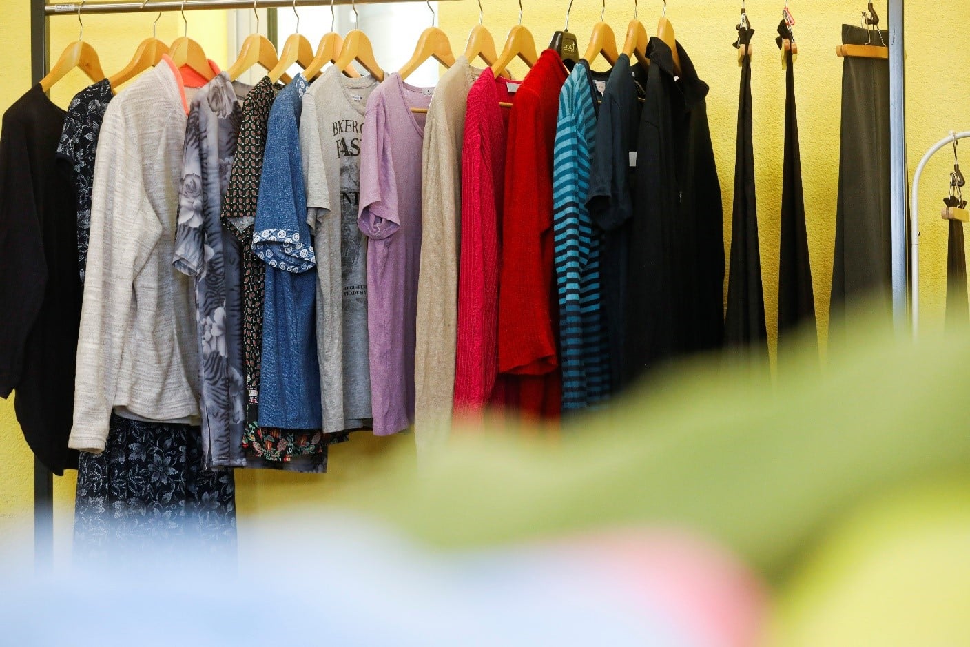 Eine Reihe verschiedener Hemden, Kleider und Oberteile in unterschiedlichen Farben und Mustern hängt auf Holzbügeln vor einer gelben Wand.