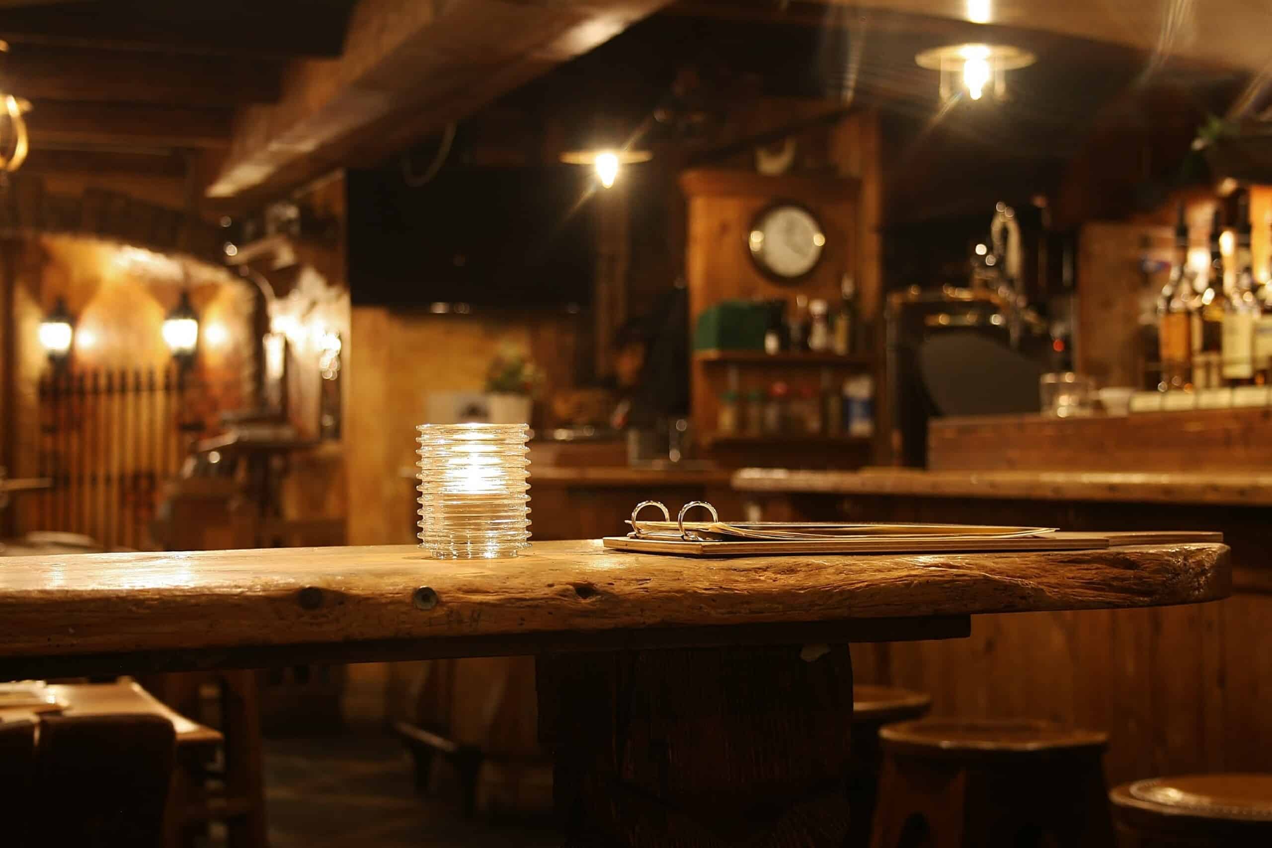 Ein Holztisch in einer schwach beleuchteten Bar mit einem Stapel durchsichtiger Untersetzer und einer geschlossenen Speisekarte; im Hintergrund sind Regale mit Flaschen zu sehen.