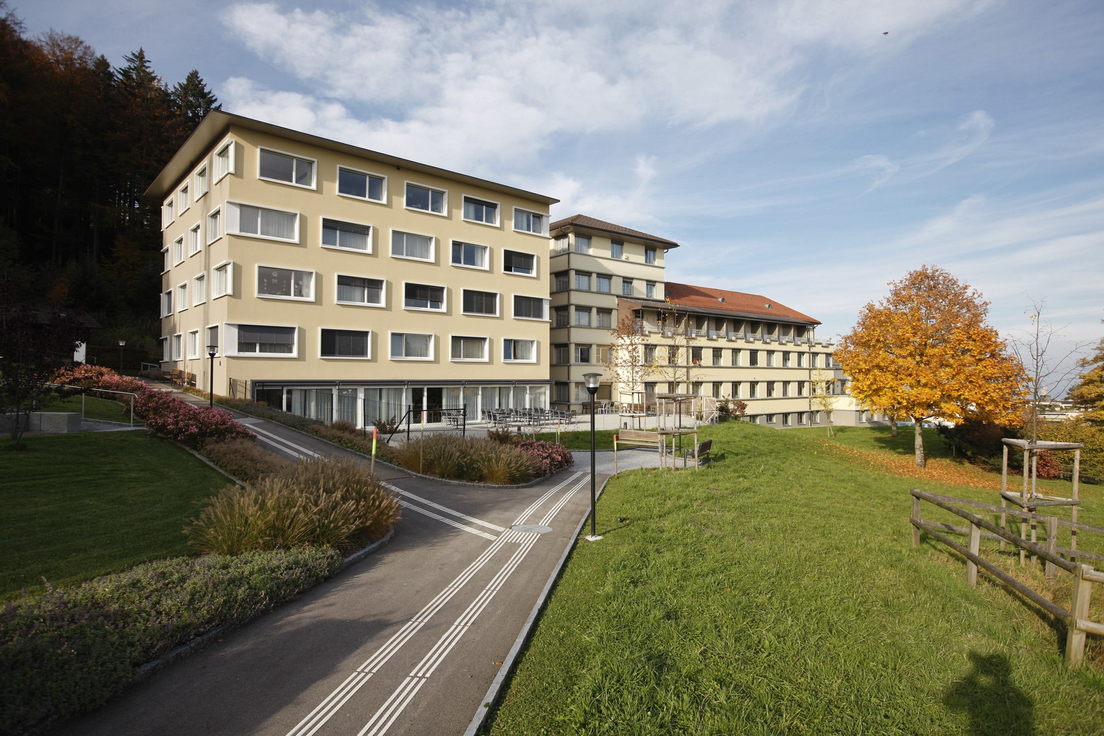 Ein modernes mehrstöckiges Gebäude mit beiger Fassade und vielen Fenstern, umgeben von Grün, Wegen und Herbstbäumen unter einem teilweise bewölkten Himmel.