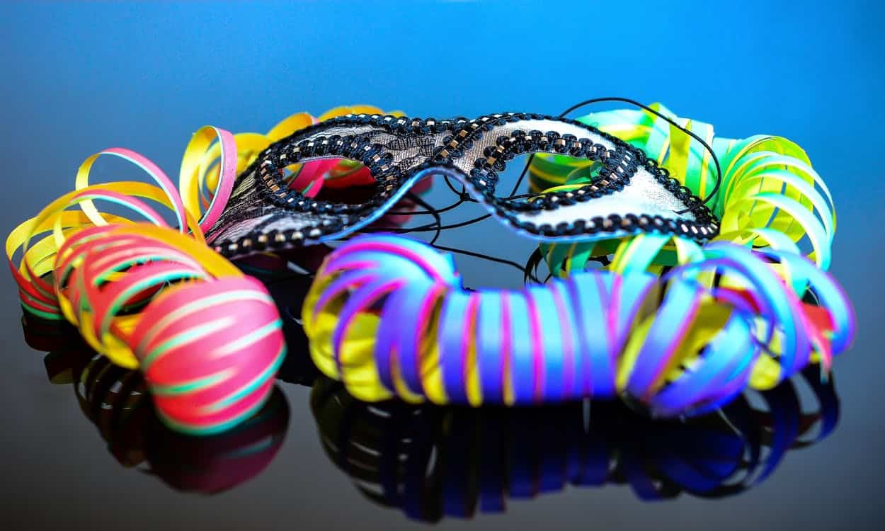 Eine schwarz-weiße Maskenmaske liegt auf einer reflektierenden Oberfläche, umgeben von bunten, gewellten Bändern unter blauem Licht.