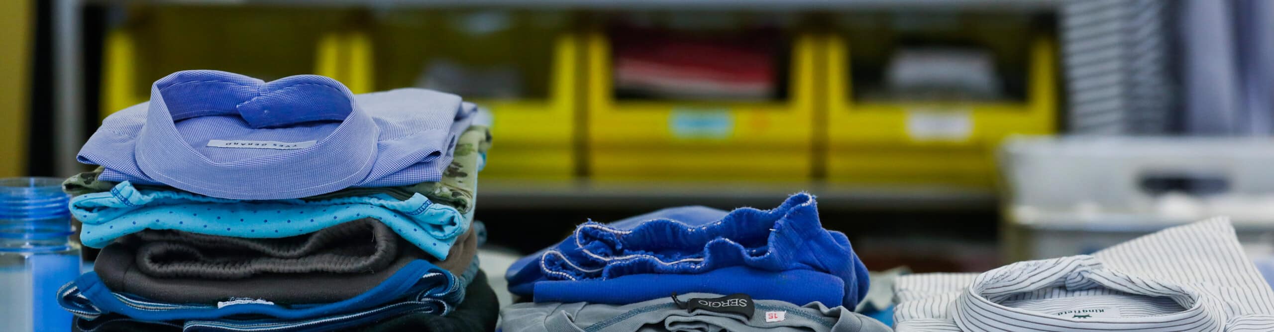 Stapel gefalteter Hemden in verschiedenen Blau- und Grautönen sind auf einem Tisch angeordnet, im Hintergrund stehen gelbe Lagerbehälter.