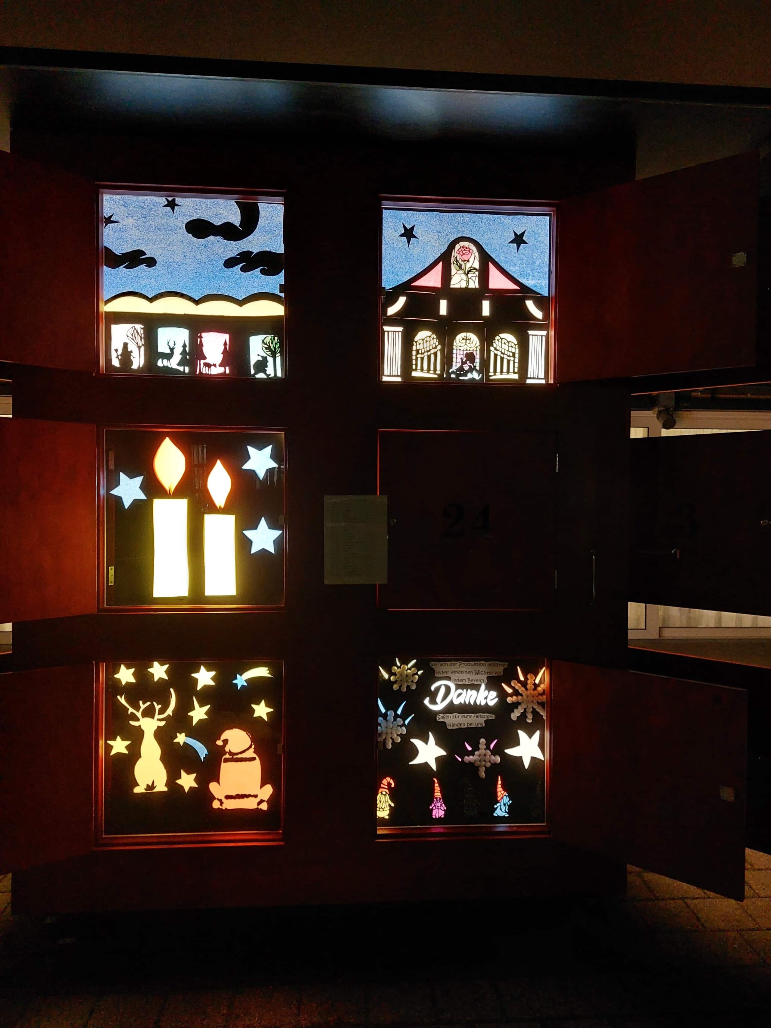 Ein Holzschrank mit beleuchteten, bunten Fensterscheiben, die Kerzen, Sterne, Figuren und festliche Szenen darstellen, wahrscheinlich Teil einer Adventskalender-Ausstellung.