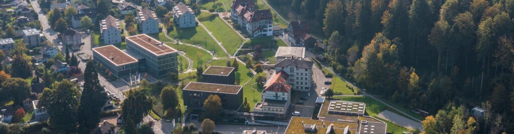 Luftaufnahme eines Vorortgebiets mit modernen Gebäuden, Wohnhäusern, Gründächern und angrenzendem Waldgebiet.