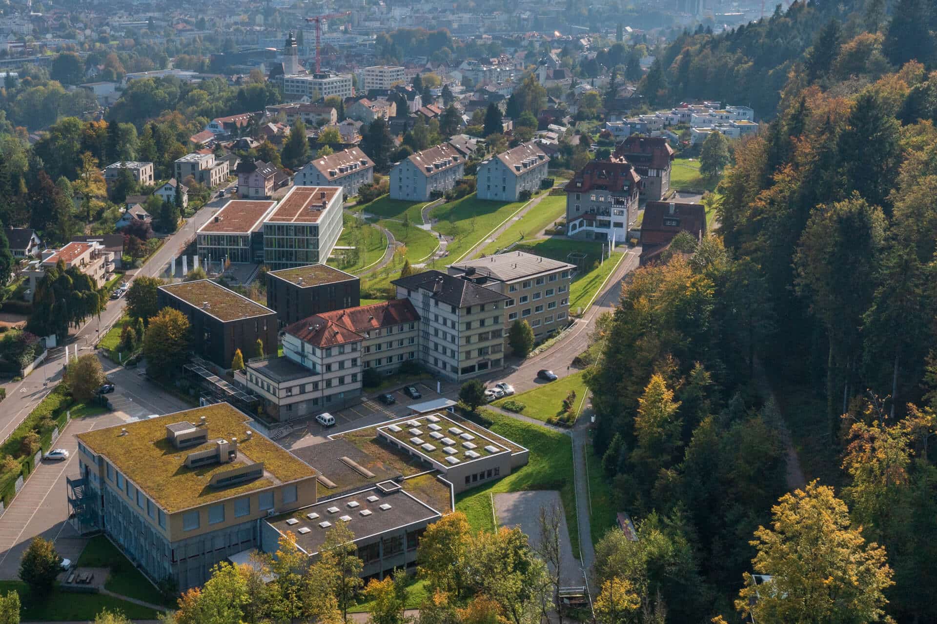 Luftaufnahme einer Kleinstadt mit modernen und traditionellen Gebäuden, Straßen, Grünflächen und einem bewaldeten Hügel auf der rechten Seite.
