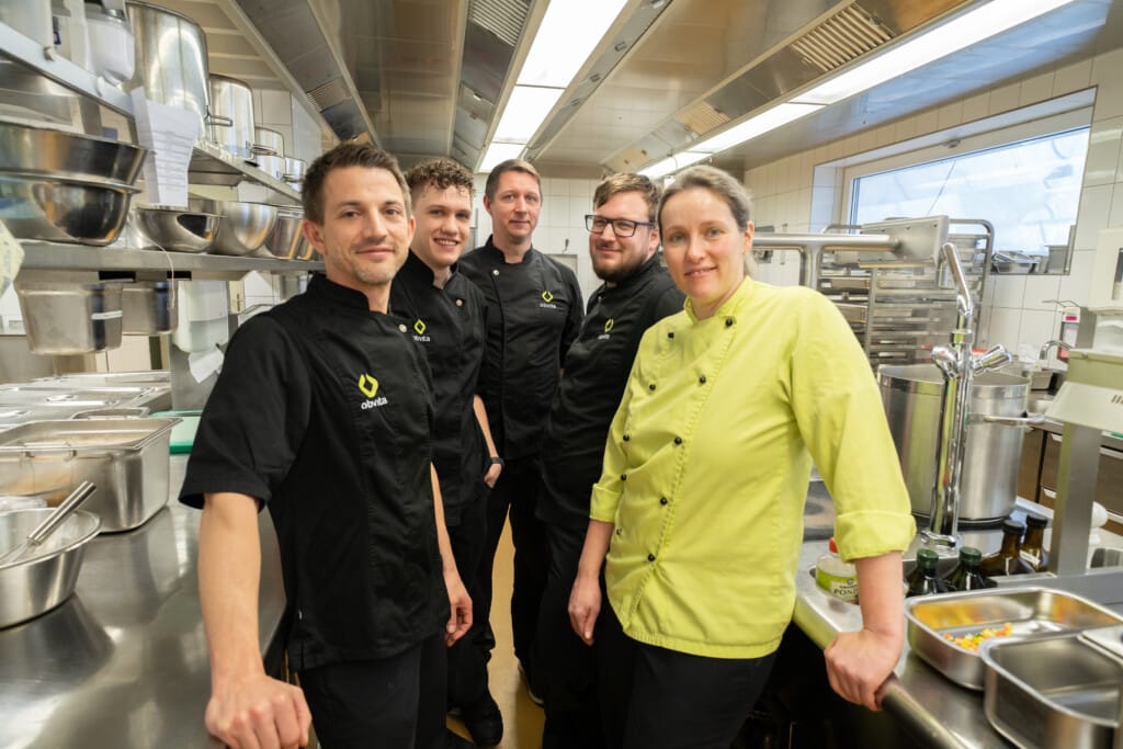 Fünf Köche in Uniform stehen zusammen in einer Großküche, umgeben von Geräten und Theken aus Edelstahl.