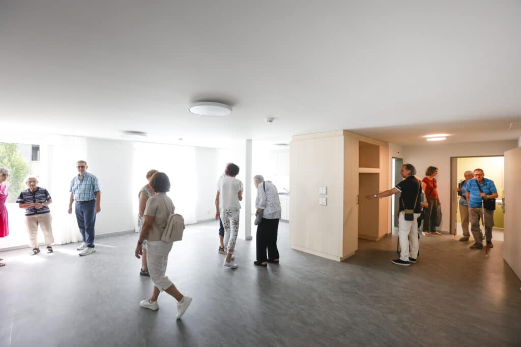 Eine Gruppe von Menschen besichtigt einen hellen, unmöblierten Raum mit großen Fenstern und hellen Wänden und Böden.