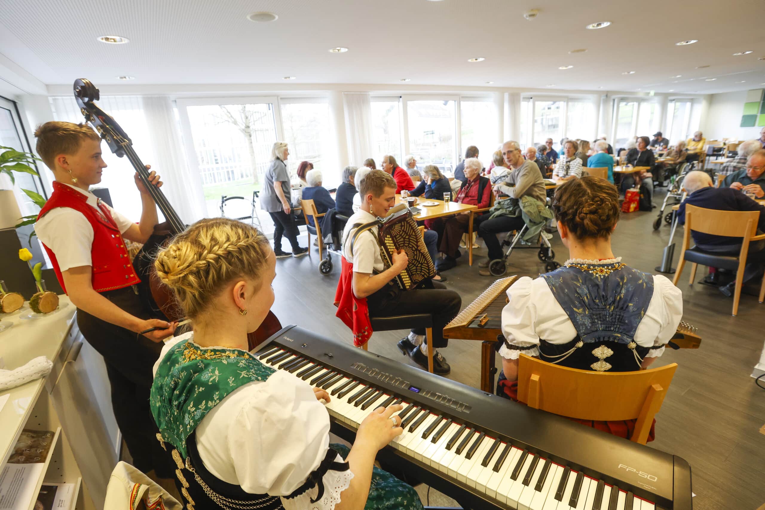Eine kleine Gruppe junger Musiker in traditioneller Kleidung spielt auf Keyboard, Bass, Akkordeon und Hackbrett vor Menschen, die in einem hell erleuchteten, belebten Café oder Speisesaal sitzen.