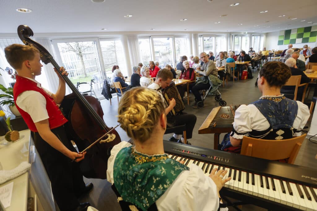 In einem hellen, modernen Raum mit großen Fenstern treten Musiker in traditioneller Kleidung vor einem sitzenden Publikum auf.