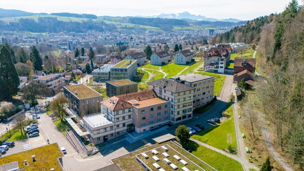 Luftaufnahme eines Wohngebiets am Hang mit Wohnhäusern, grünen Rasenflächen, Bäumen und einer Stadtlandschaft im Hintergrund.