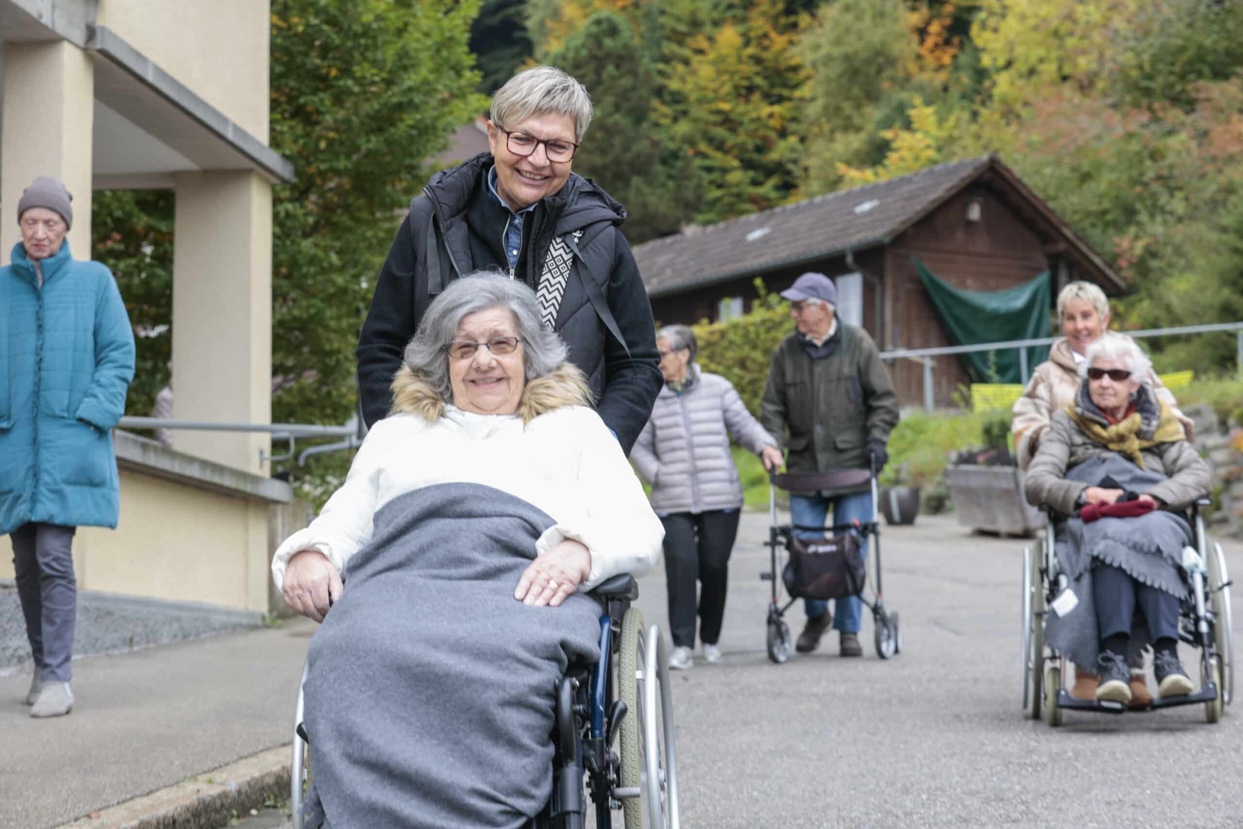 Eine Frau schiebt eine ältere Frau im Rollstuhl nach draußen, während mehrere andere Personen, einige mit Rollatoren, in der Nähe auf einem gepflasterten Weg gehen. Im Hintergrund sind Bäume und ein Holzgebäude zu sehen.